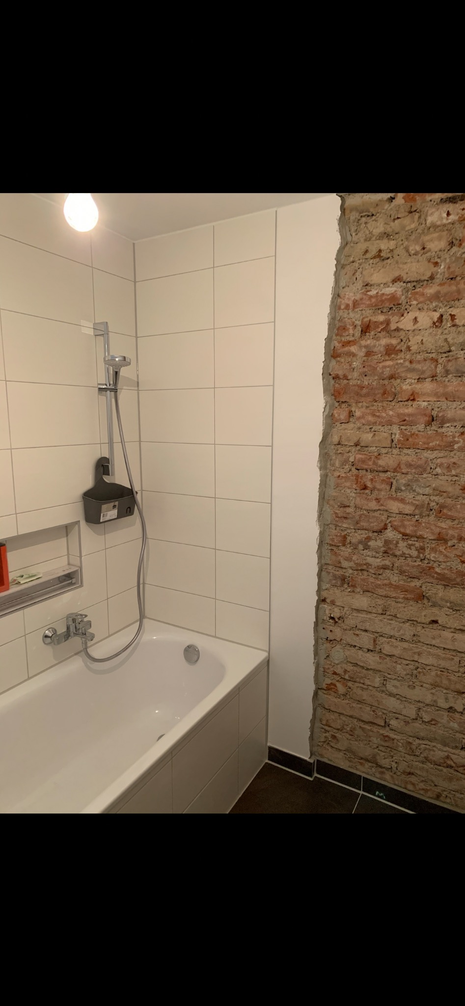 Biała wanna z baterią i prysznicem na tle białych płytek. Fragment ściany z surowej cegły. Kontrastowe wykończenie podłogi. Etap prac w łazience.