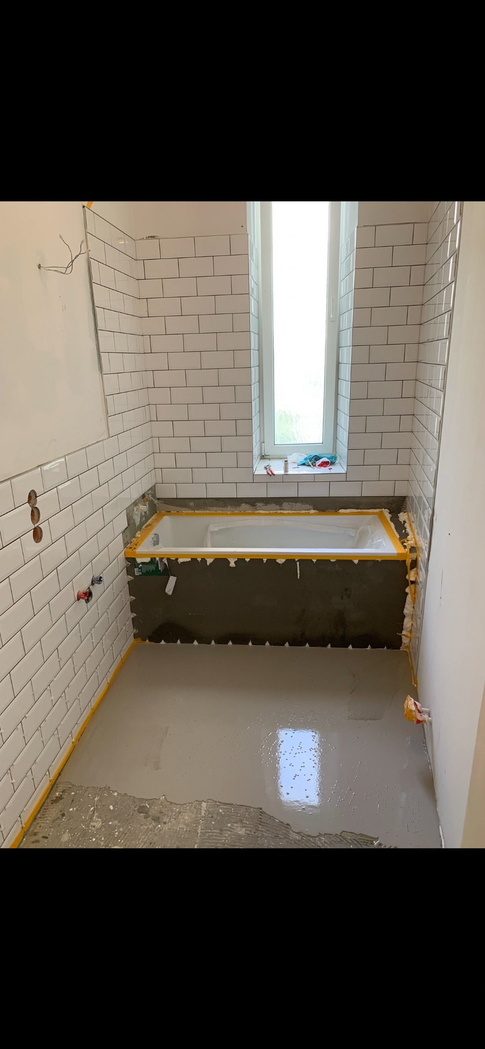 Łazienka w trakcie remontu: białe płytki typu metro na ścianach, przygotowana wanna z taśmą zabezpieczającą i wylewka na podłodze. Widoczne niedokończone prace.