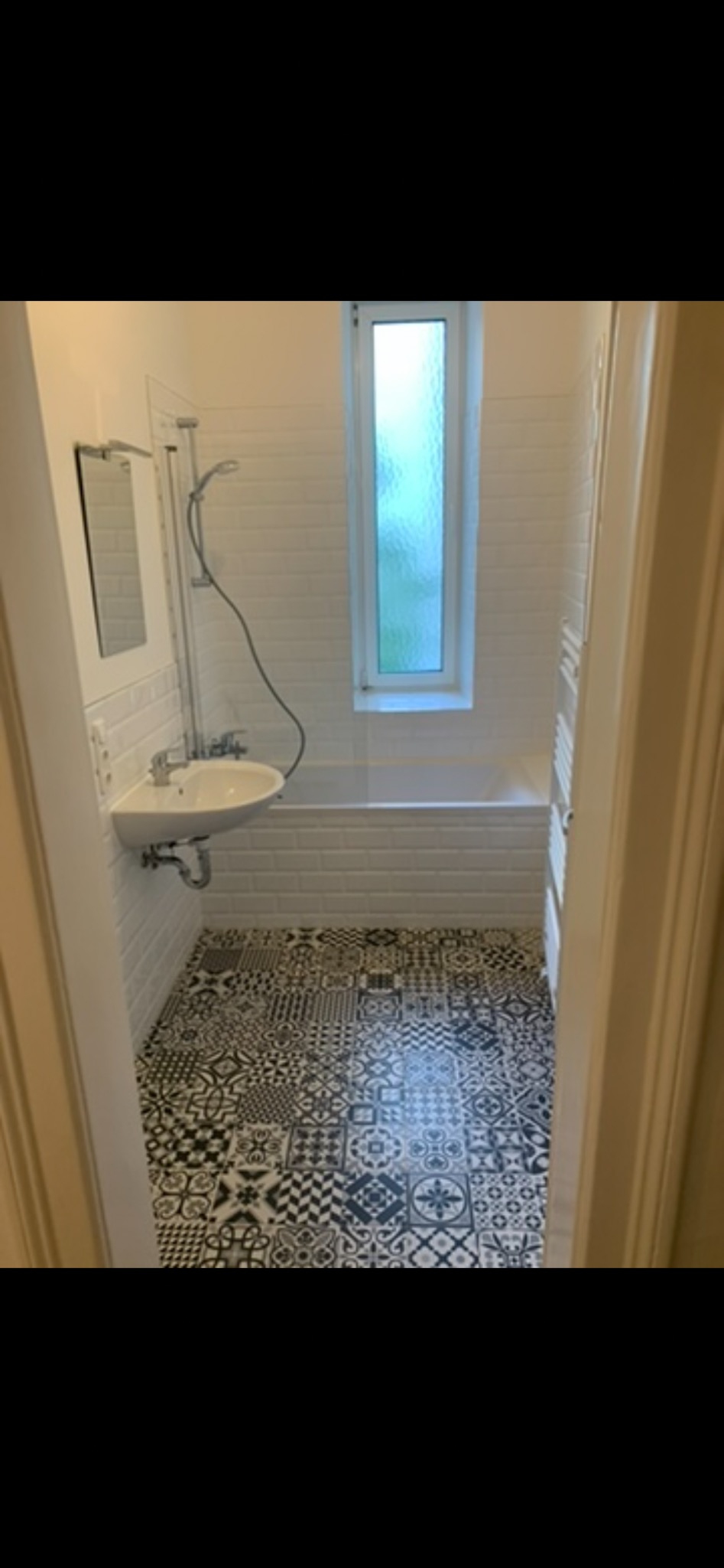 Wykończona łazienka w Iławie z wanną, umywalką i wzorzystą podłogą. Białe płytki na ścianach i wąskie okno nad wanną. Widok zza otwartych drzwi.