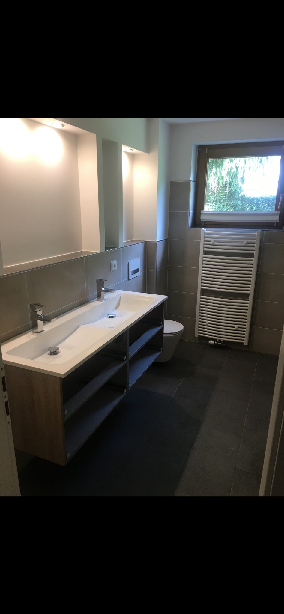 Nowoczesna łazienka z podwójną umywalką, szarą glazurą i grzejnikiem drabinkowym przy oknie. Minimalistyczny design, neutralna kolorystyka, oświetlenie punktowe.