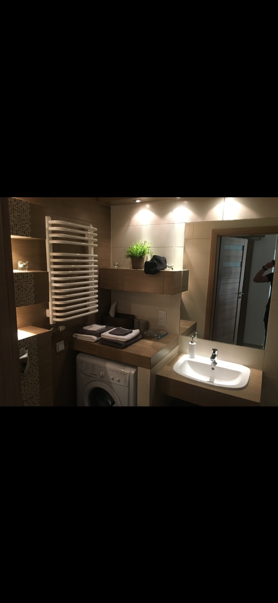Jasna łazienka z pralką pod blatem, grzejnikiem drabinkowym i dużym lustrem. Umywalka nablatowa, beżowe płytki i drewniane akcenty tworzą przytulne wnętrze.