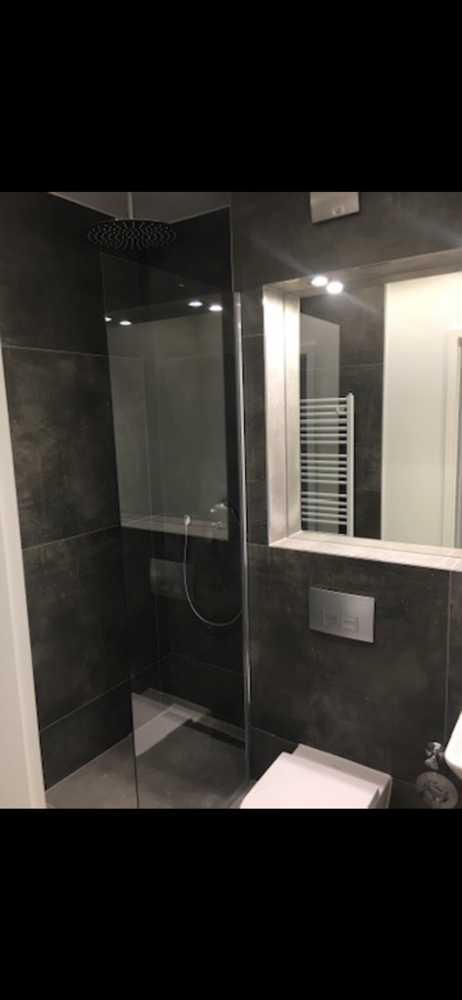 Nowoczesna łazienka z kabiną typu walk-in, wykończona ciemnymi płytkami imitującymi kamień. Widoczny deszczownica, lustro z oświetleniem i grzejnik drabinkowy.