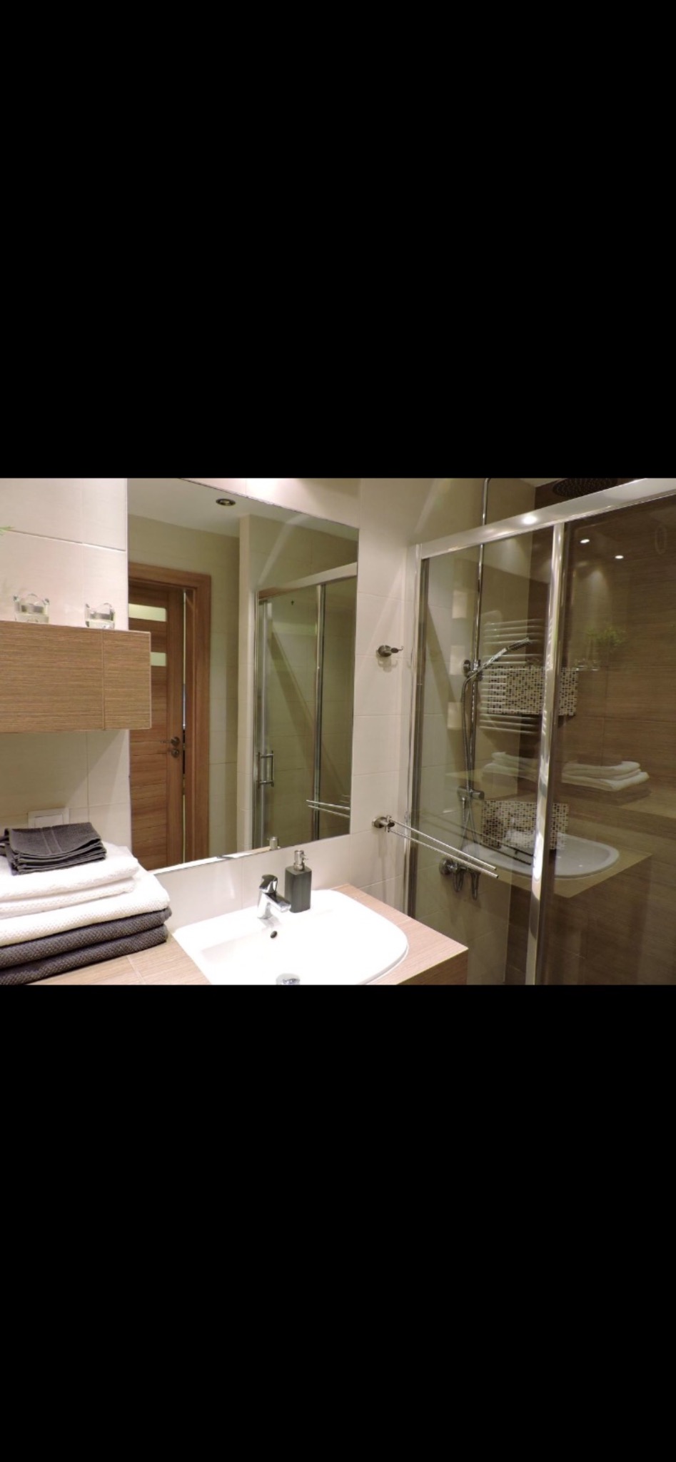 Nowoczesna łazienka z kabiną prysznicową, umywalką i stertą ręczników. Jasne płytki i drewniane akcenty tworzą przytulne wnętrze. Widoczne detale wykończenia.