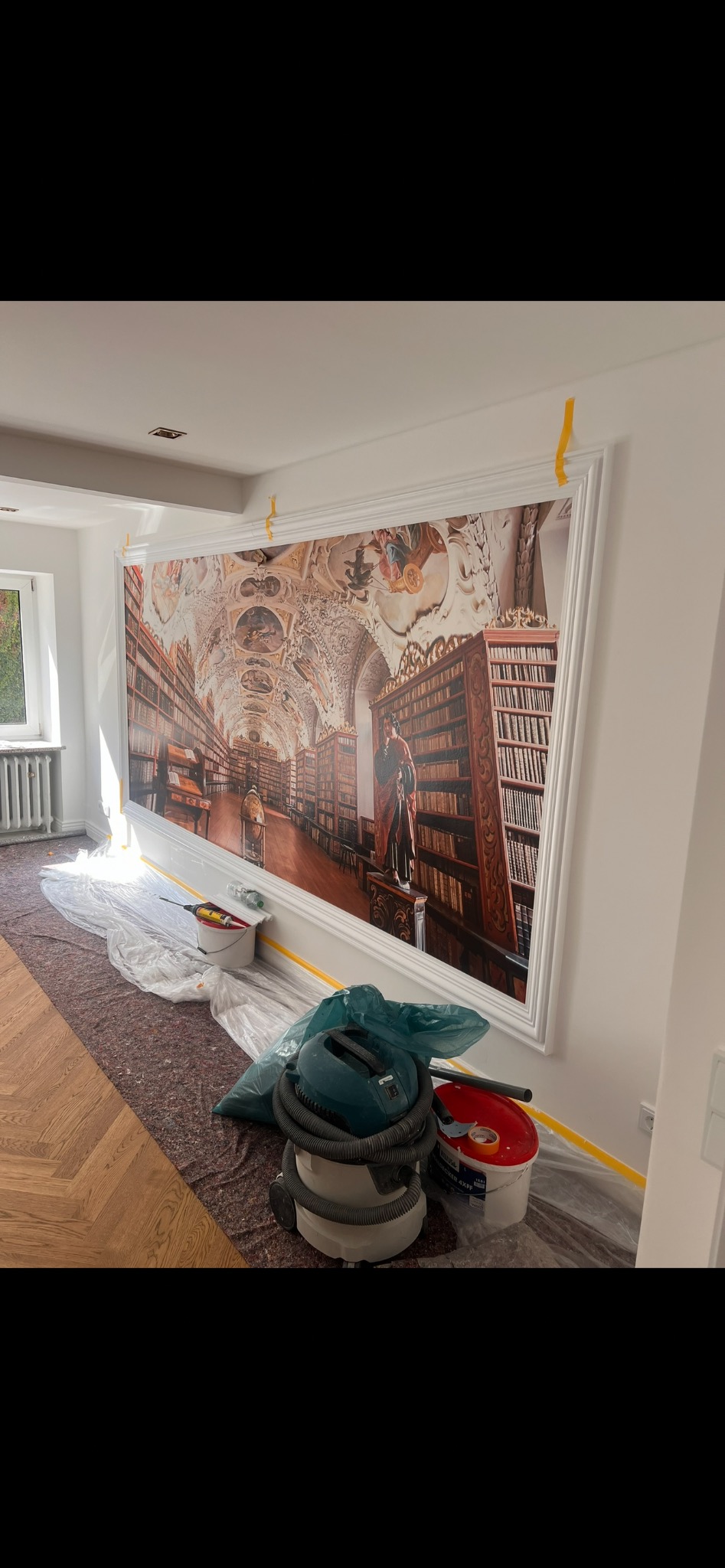 Montaż fototapety z motywem biblioteki w ozdobnej ramie. Podłoga zabezpieczona folią, widoczny odkurzacz i wiadro z farbą. Żółta taśma malarska na ścianach.