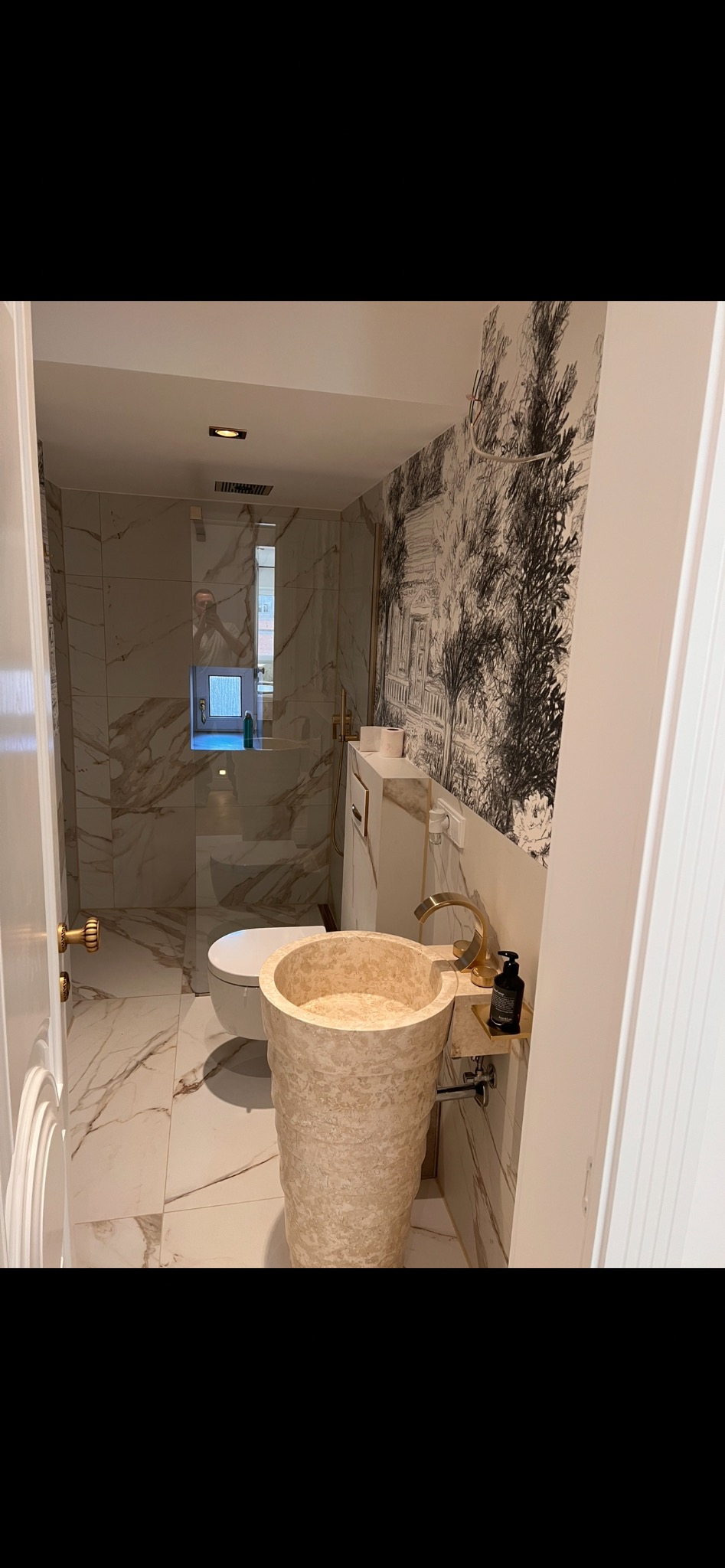 Elegancka łazienka z marmurowymi płytkami, wolnostojącą umywalką z trawertynu i dekoracyjną tapetą w stylu vintage. Widoczna kabina prysznicowa i złote akcenty.