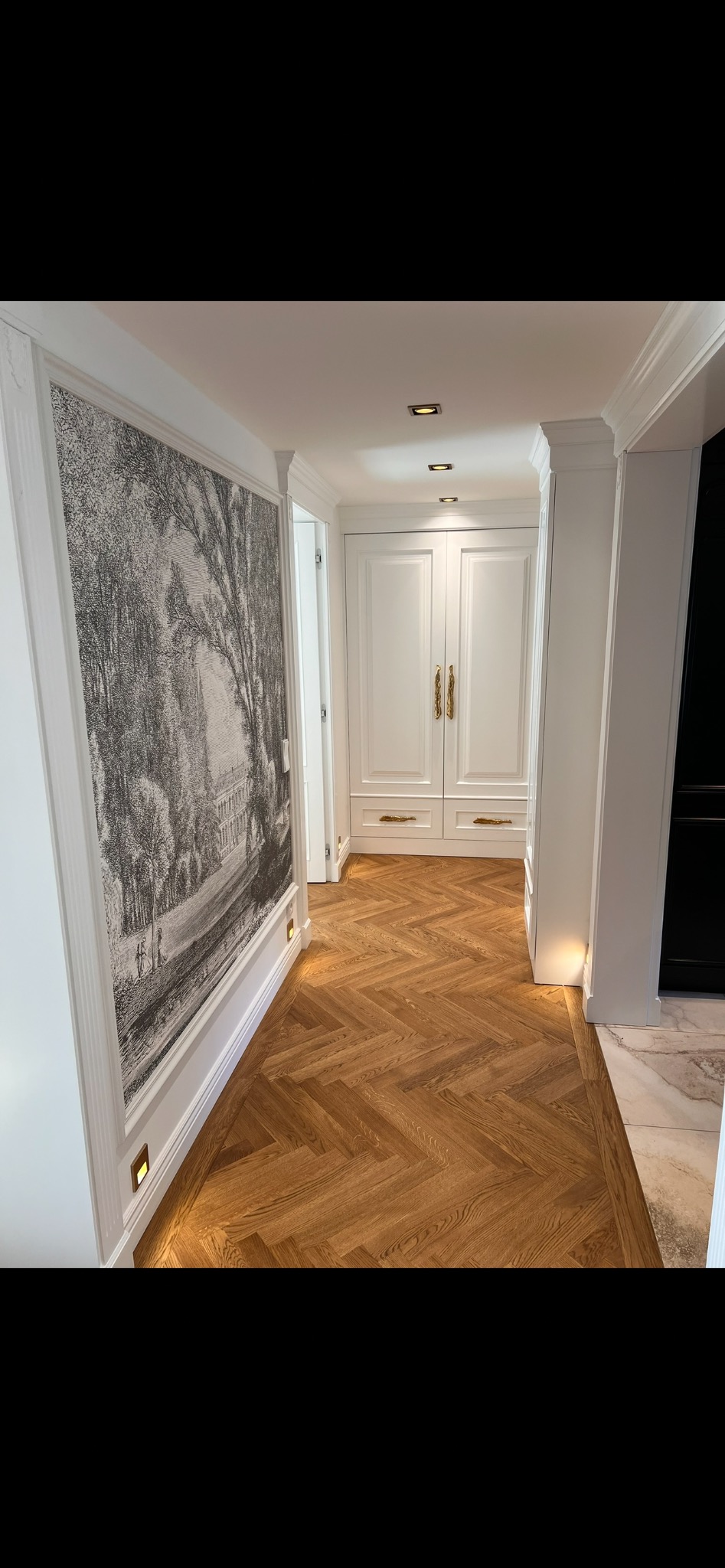 Elegancki korytarz z parkietem w jodełkę, białą zabudową i dekoracyjną grafiką na ścianie. Oświetlenie punktowe podkreśla detale wykończenia.