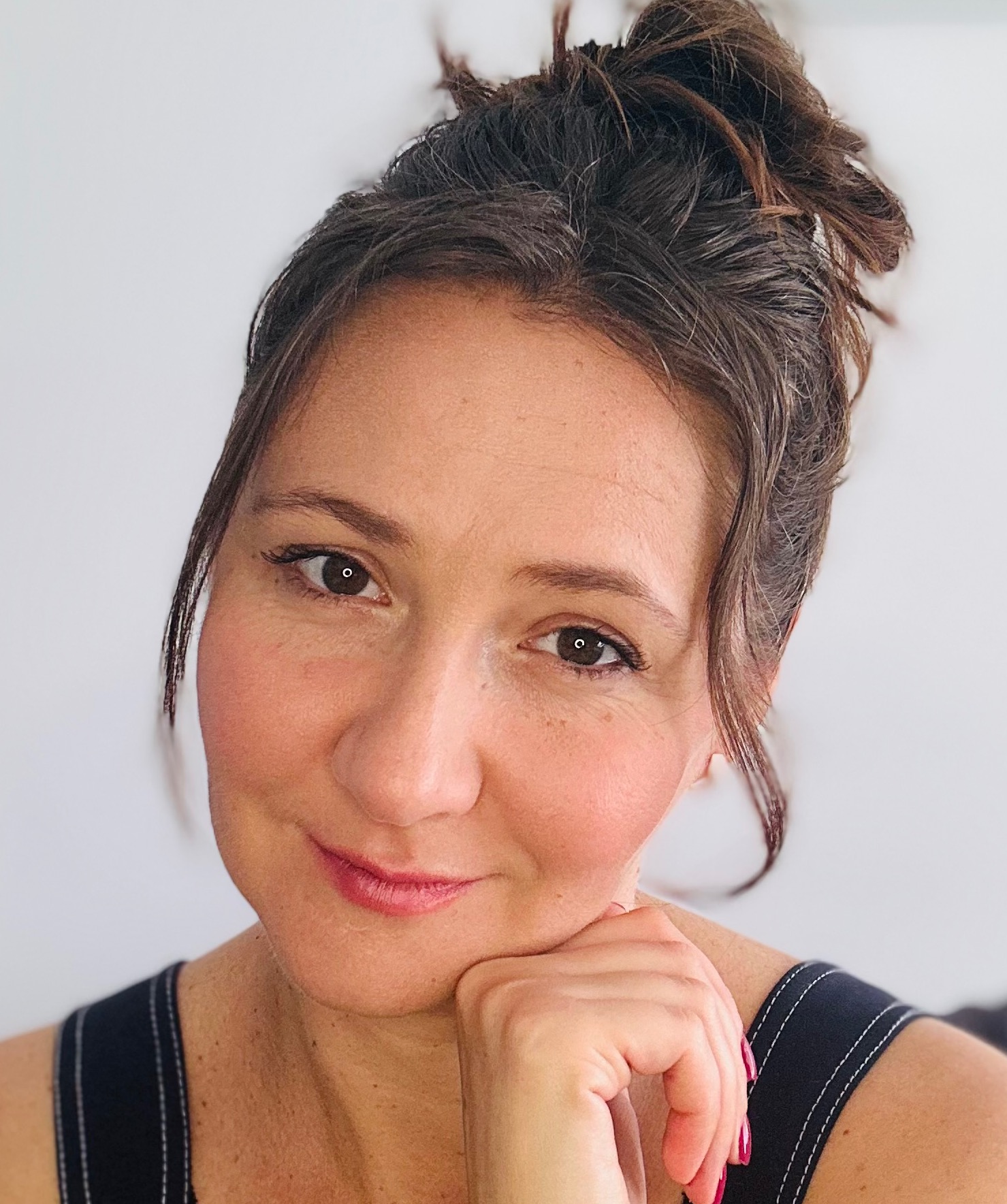 Portret uśmiechniętej kobiety z brązowymi włosami upiętymi w kok, opierającej podbródek na dłoni. Widoczne delikatne zmarszczki mimiczne i różowe paznokcie.