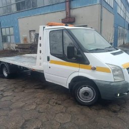 Orlov Auto Service - Biała laweta Ford Transit na mokrym bruku przed budynkiem przemysłowym. Pomarańczowe światła ostrzegawcze na dachu. Widok z boku, pochmurny dzień.