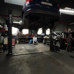 Orlov Auto Service - Niebieski Ford Focus na podnośniku w warsztacie samochodowym. Widoczny mechanik i narzędzia. Oświetlenie jarzeniowe, szara podłoga.