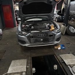 Orlov Auto Service - Srebrne Audi A6 z otwartą maską w warsztacie samochodowym, widoczny silnik i skrzynka z częściami. W tle podnośnik i inne pojazdy. Ujęcie z kanału.