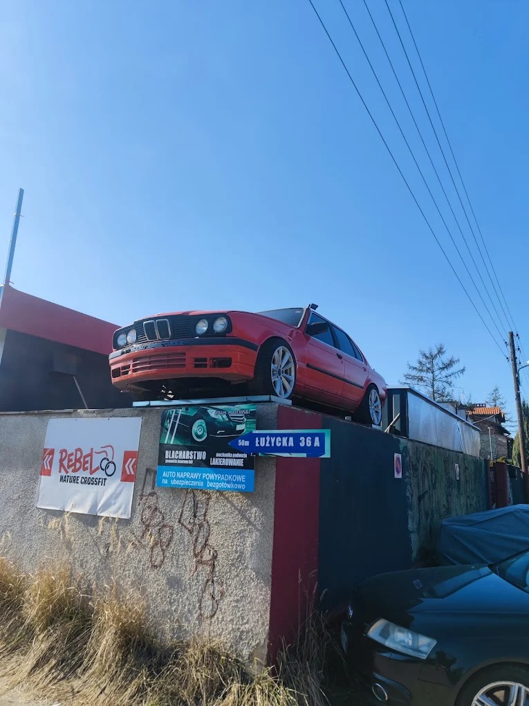 Czerwone BMW E30 umieszczone na podwyższeniu, reklama warsztatu blacharsko-lakierniczego z adresem 'Łużycka 36A'. Nietypowa ekspozycja auta.