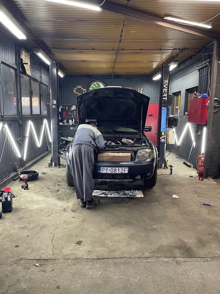 Mechanik w warsztacie samochodowym pochylony nad silnikiem auta z otwartą maską. Widoczne narzędzia i oświetlenie warsztatowe.