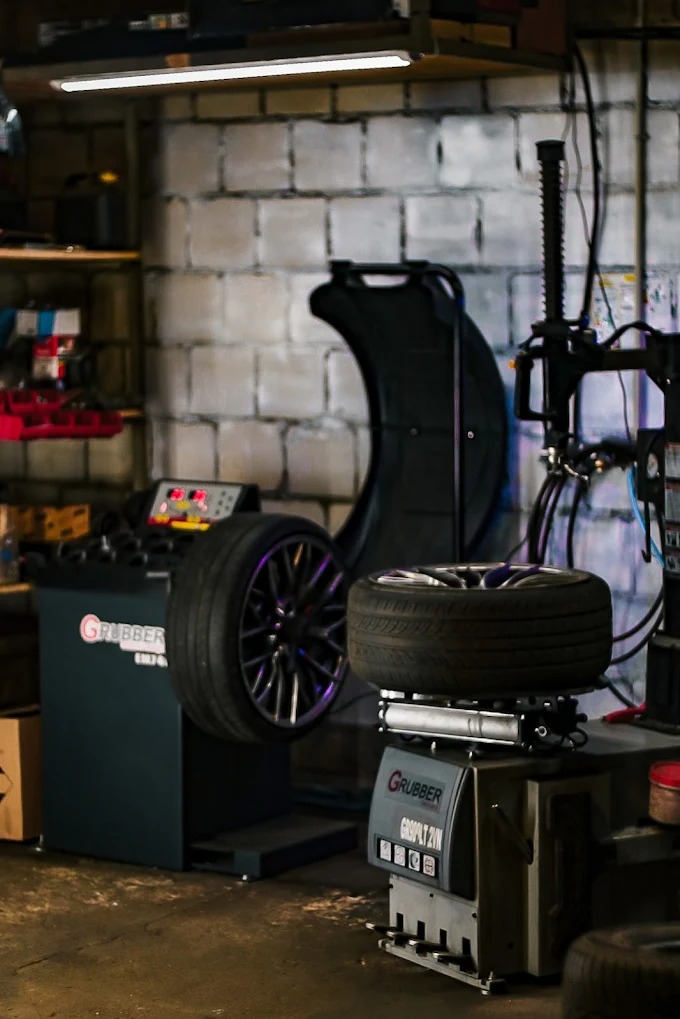 Wyważarka do kół w warsztacie. Dwa koła na maszynach, ceglana ściana w tle. Sprzęt warsztatowy marki Grubber. Oświetlenie jarzeniowe.