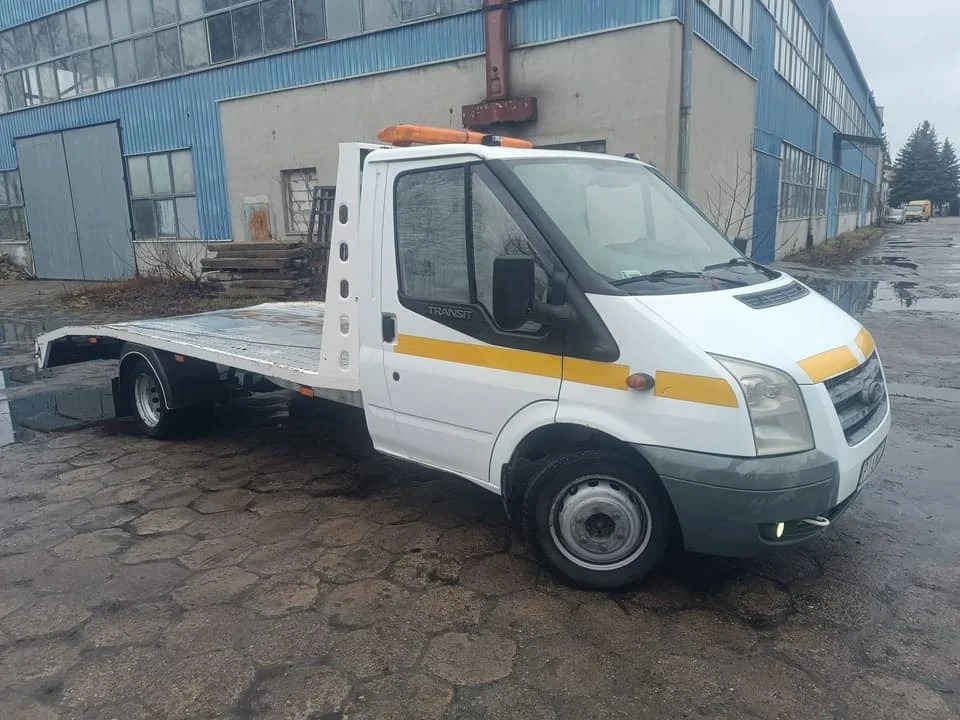 Biała laweta Ford Transit na mokrym bruku przed budynkiem przemysłowym. Pomarańczowe światła ostrzegawcze na dachu. Widok z boku, pochmurny dzień.