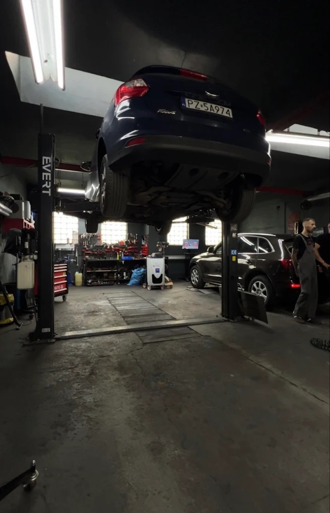 Niebieski Ford Focus na podnośniku w warsztacie samochodowym. Widoczny mechanik i narzędzia. Oświetlenie jarzeniowe, szara podłoga.