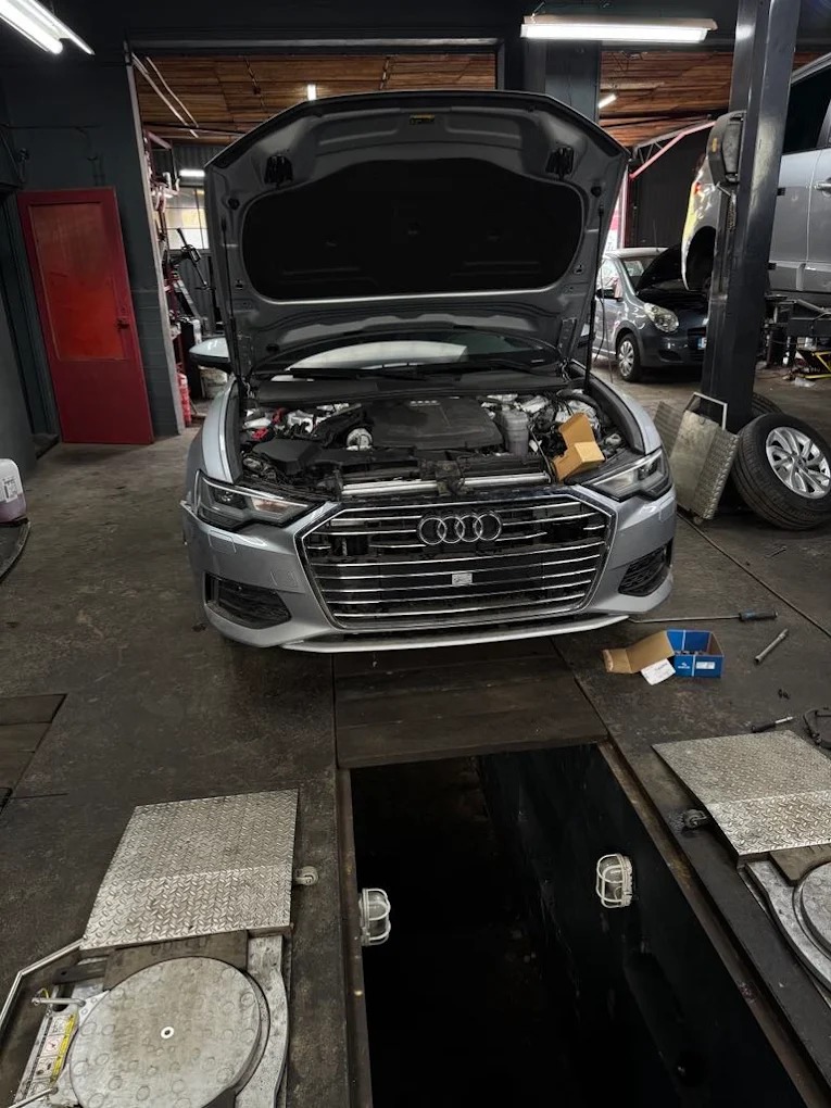 Srebrne Audi A6 z otwartą maską w warsztacie samochodowym, widoczny silnik i skrzynka z częściami. W tle podnośnik i inne pojazdy. Ujęcie z kanału.