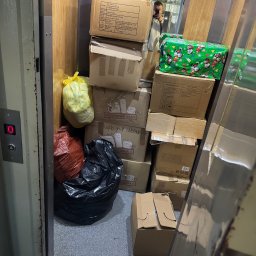 OpróżnijTo - Przepełniona winda z kartonami i workami podczas przeprowadzki. Widoczne lustro z odbiciem osoby robiącej zdjęcie. Kartony z napisem 'Przeprowadzka'.