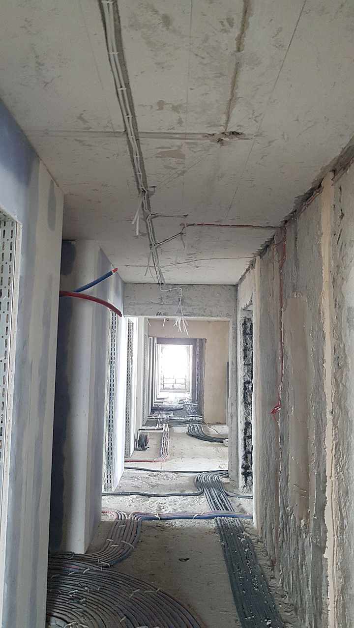 Korytarz w trakcie remontu z widocznymi instalacjami elektrycznymi na suficie i podłodze. Surowy stan wykończenia ścian i podłóg. Widok w głąb korytarza.