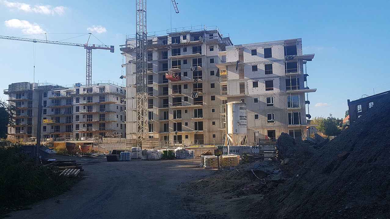 Wielopiętrowy budynek mieszkalny w trakcie budowy, widoczne surowe ściany, okna bez szyb, żurawie budowlane w tle, materiały budowlane na placu.