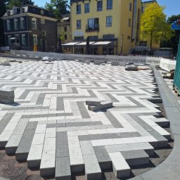 AAA INVEST SPÓŁKA Z OGRANICZONĄ ODPOWIEDZIALNOŚCIĄ - Układanie kostki brukowej w zygzak z szarej i białej kostki. Częściowo ułożona nawierzchnia na tle budynków i barierki. Widoczne niedokończone fragmenty.