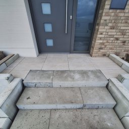 AAA INVEST SPÓŁKA Z OGRANICZONĄ ODPOWIEDZIALNOŚCIĄ - Wykończone schody wejściowe z betonowych elementów, prowadzące do nowoczesnych drzwi. Jasne płytki na podeście, ceglana ściana po prawej, koszyk dekoracyjny po lewej.