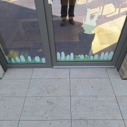 AAA INVEST SPÓŁKA Z OGRANICZONĄ ODPOWIEDZIALNOŚCIĄ - Jasne płytki tarasowe z delikatnym wzorem, widoczne przez szybę z naklejkami. Odbicie osoby w szybie. Fragment ściany z cegły po prawej stronie.