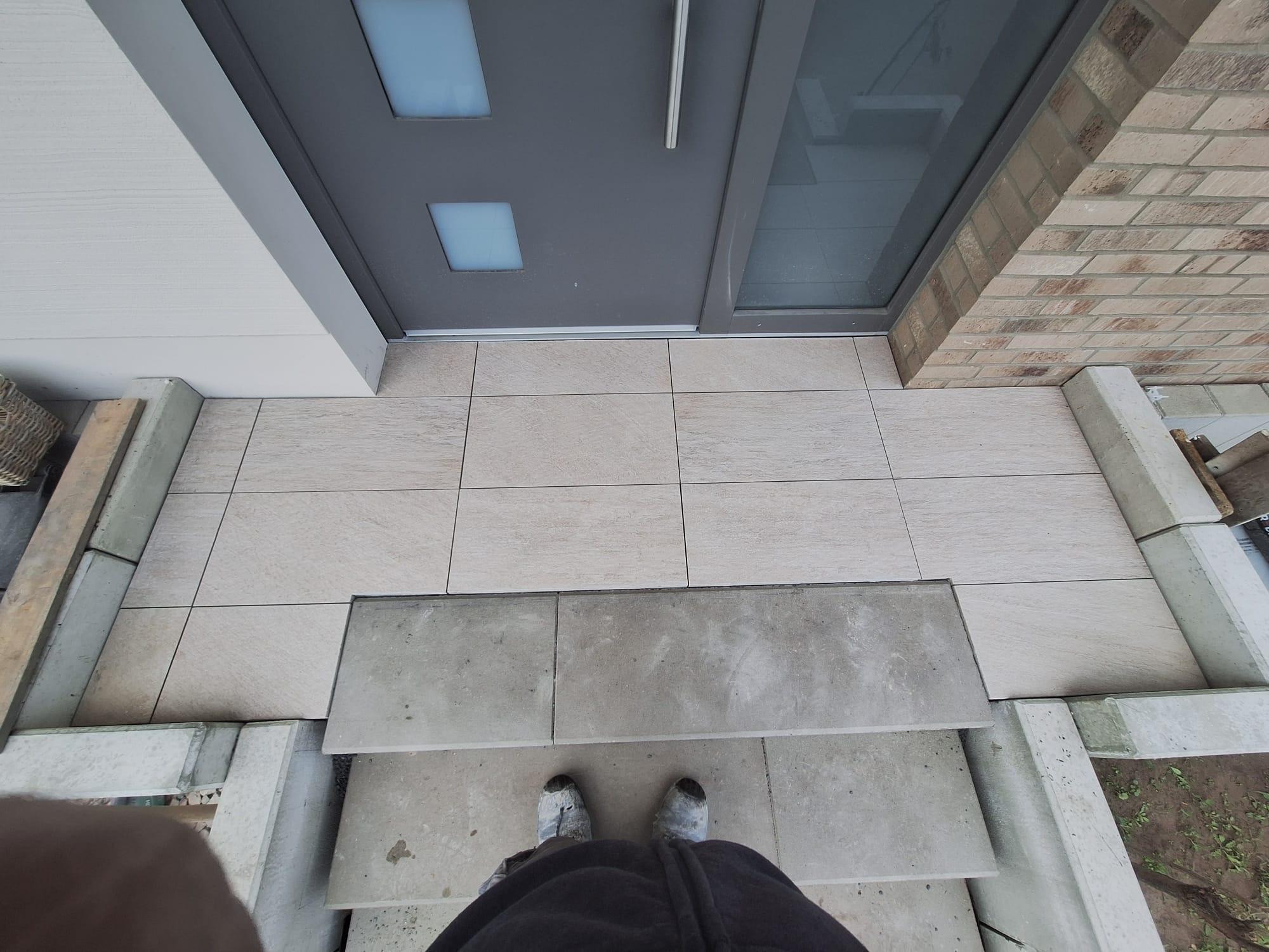 Wykończone płytki tarasowe przed wejściem do domu, z widocznymi schodami i obrzeżami. Ujęcie z góry, z butami osoby fotografującej w kadrze.