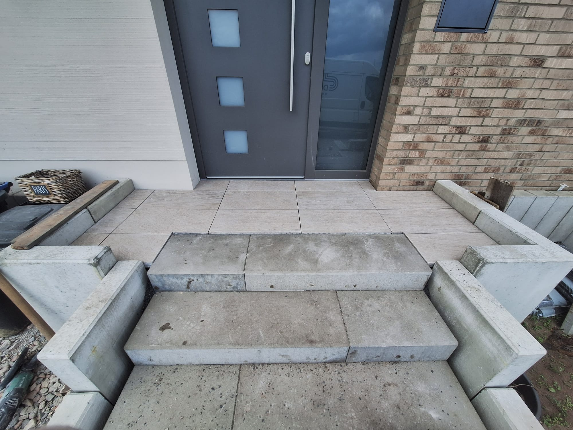 Wykończone schody wejściowe z betonowych elementów, prowadzące do nowoczesnych drzwi. Jasne płytki na podeście, ceglana ściana po prawej, koszyk dekoracyjny po lewej.