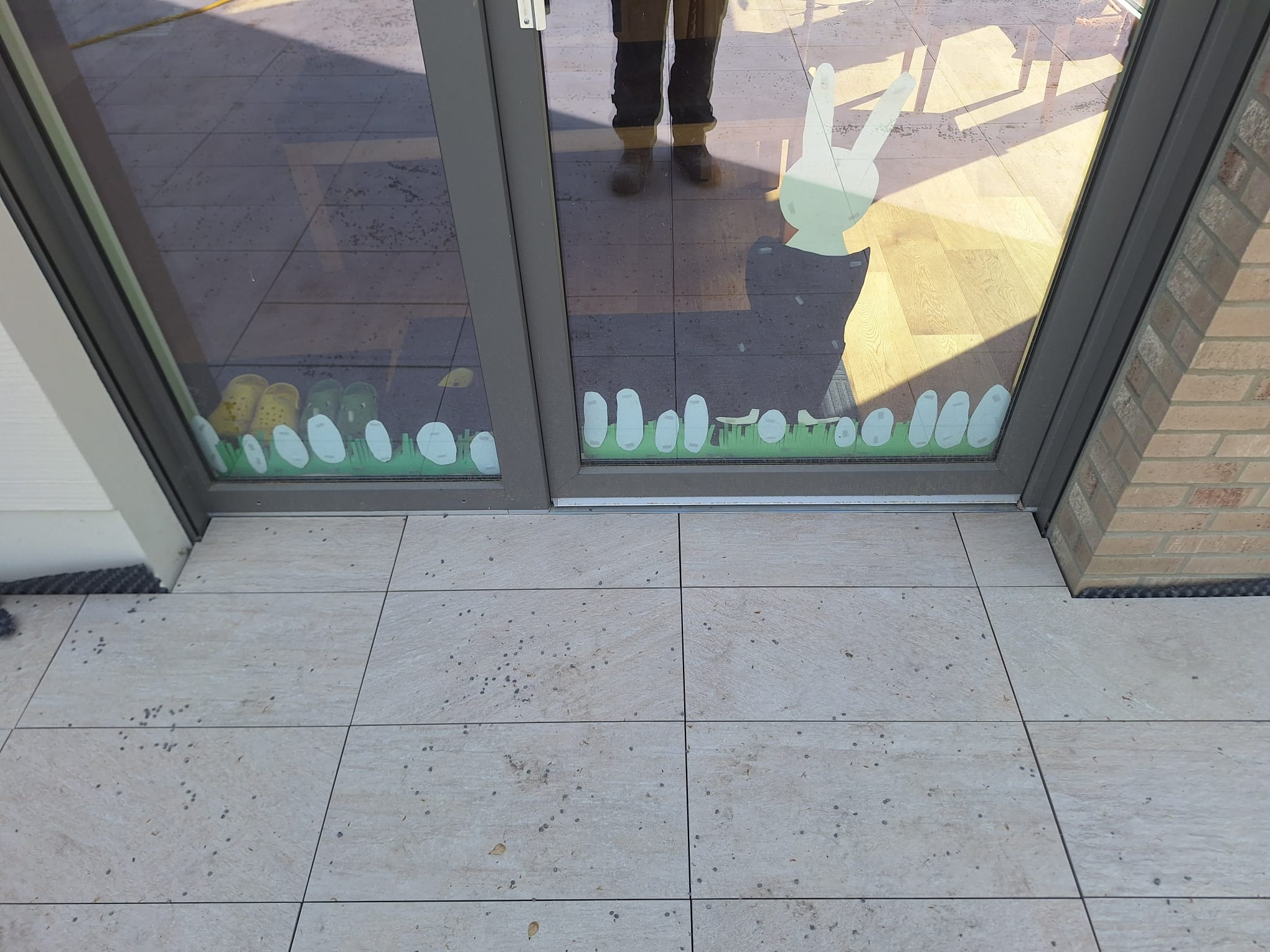 Jasne płytki tarasowe z delikatnym wzorem, widoczne przez szybę z naklejkami. Odbicie osoby w szybie. Fragment ściany z cegły po prawej stronie.