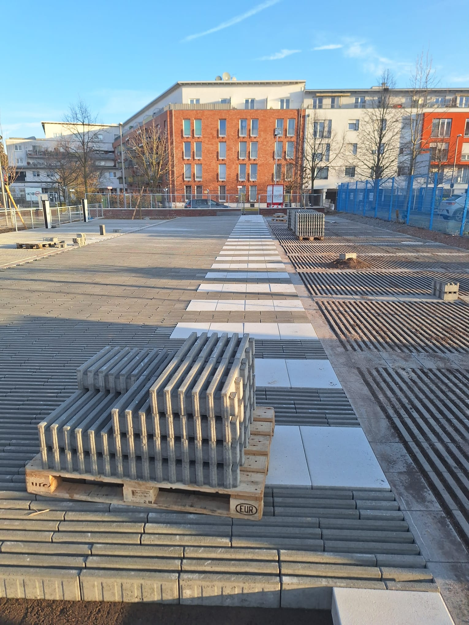 Fragment placu z ułożoną szarą kostką brukową z białymi wstawkami. Na pierwszym planie paleta z kostką. W tle budynki mieszkalne i błękitne niebo.