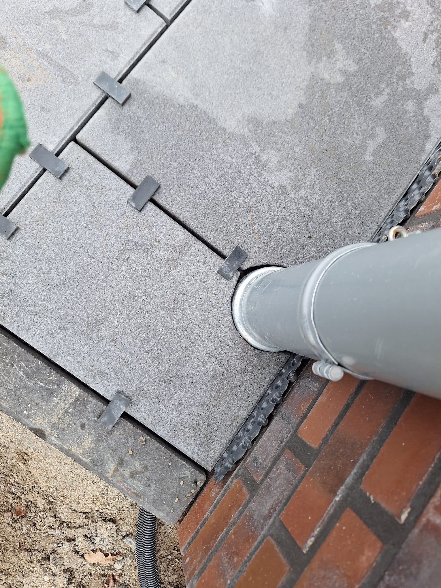 Szare płyty tarasowe z dystansami, rura odpływowa przy ceglanej ścianie. Detal montażu tarasu z odprowadzeniem wody.