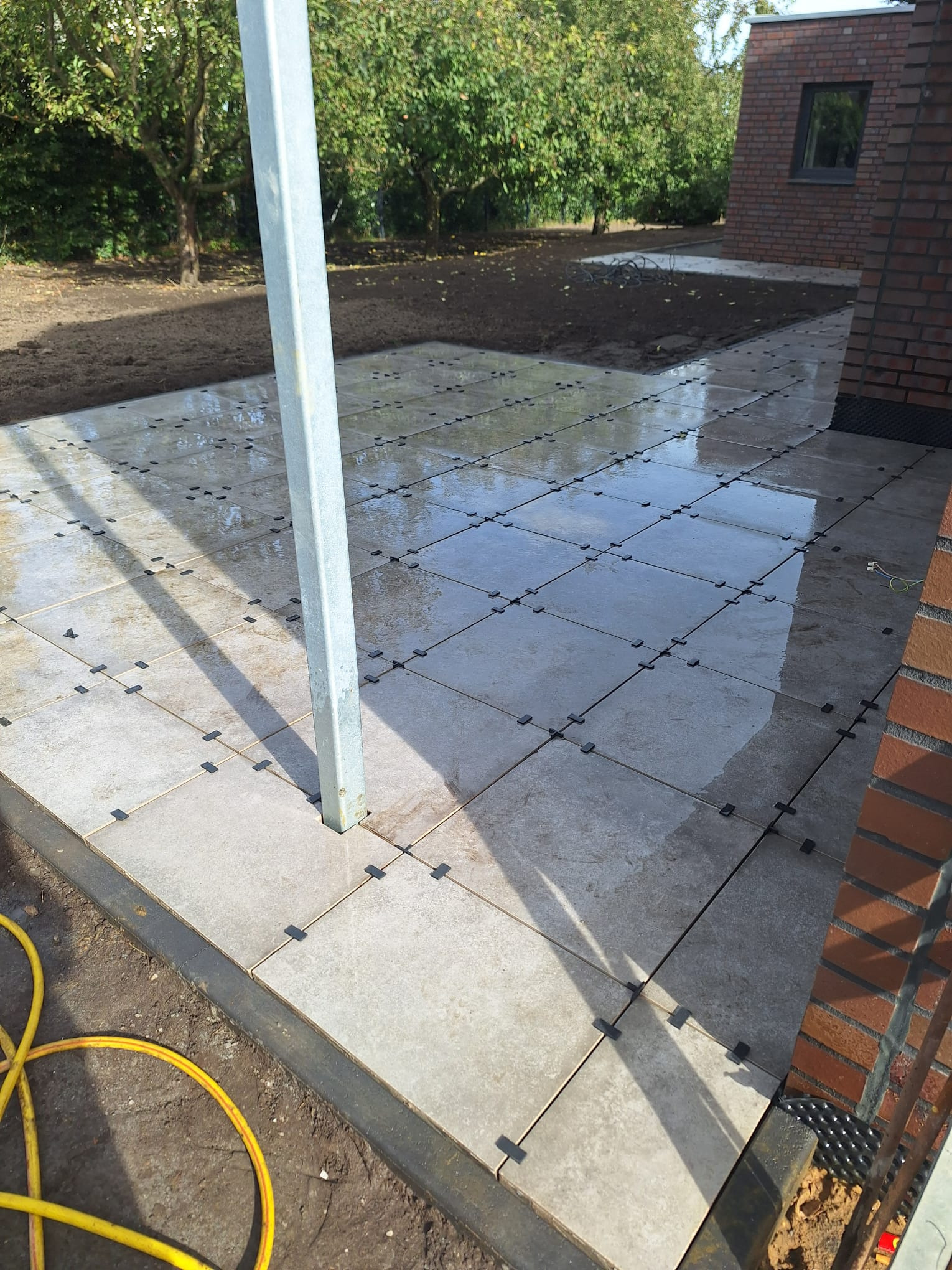 Jasne, kwadratowe płyty tarasowe ułożone na dystansach, częściowo mokre, z widocznym słupem konstrukcyjnym i fragmentem budynku z cegły w tle.