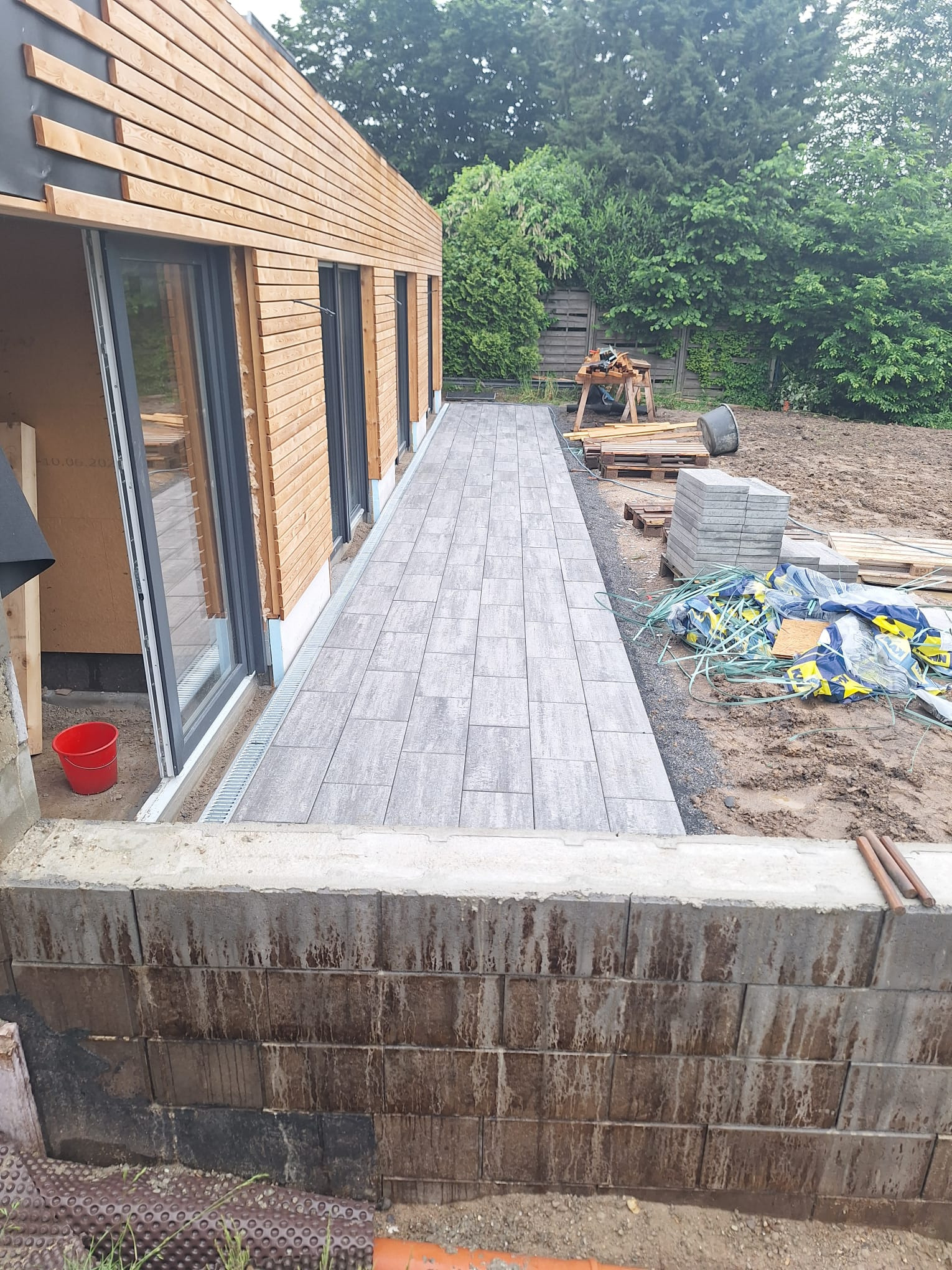 Układanie szarej kostki brukowej wzdłuż drewnianego domu z dużymi oknami. Widoczny fragment murku oporowego i przygotowany teren pod dalsze prace.
