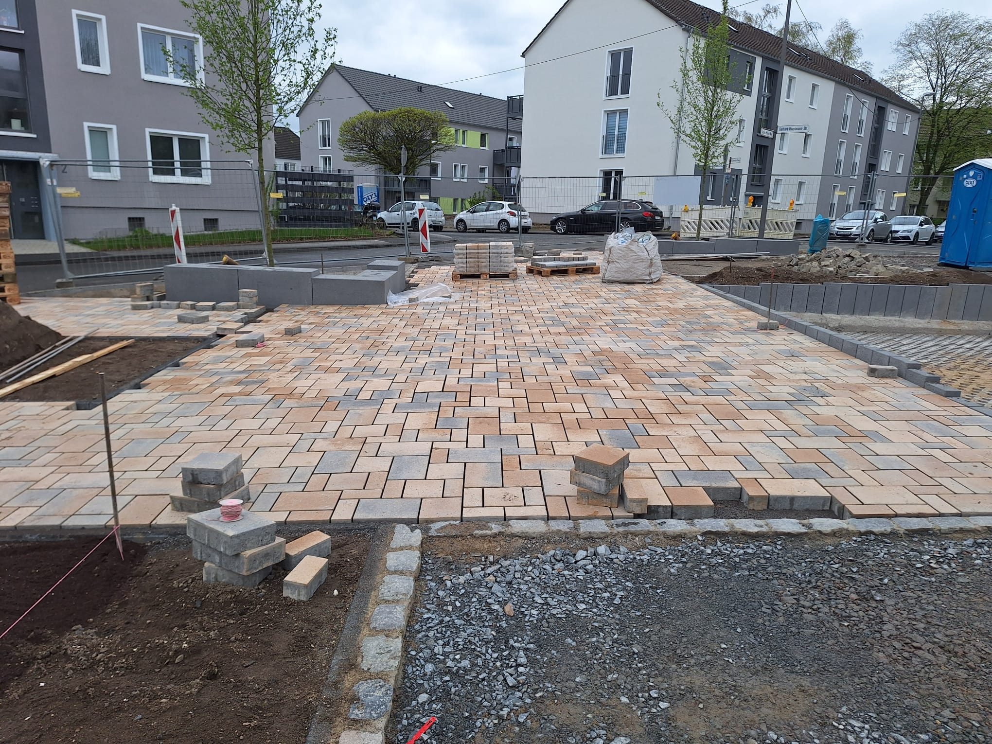 Fragment układanej nawierzchni z kostki brukowej w odcieniach beżu i szarości, z widocznymi elementami przygotowania podłoża i zapasem kostki. Praca w toku.