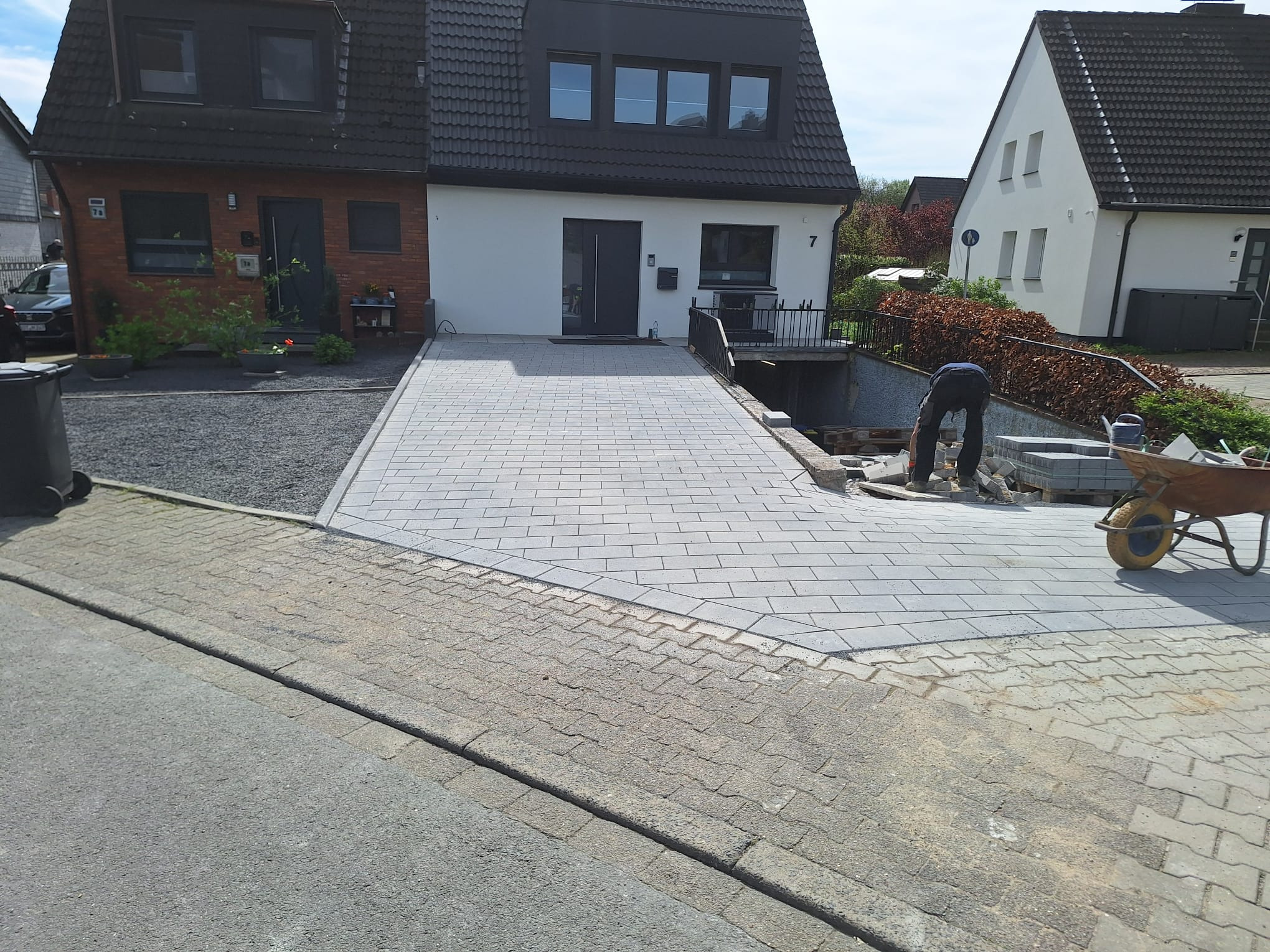 Ułożona szara kostka brukowa na podjeździe do domu, widoczny fragment ulicy i pracownik w trakcie układania, taczka z kostką obok. Dom z białymi ścianami w tle.