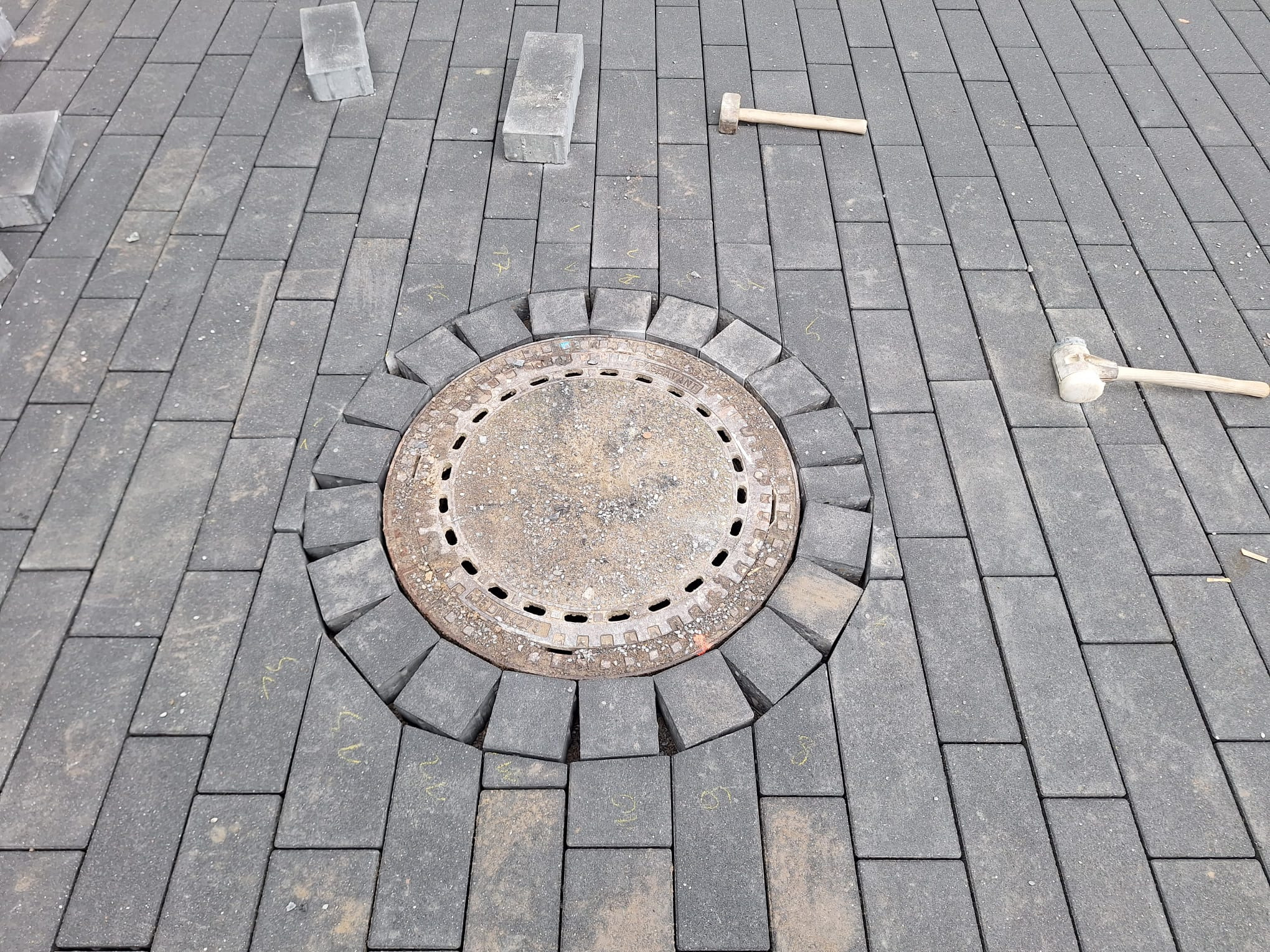 Szara kostka brukowa ułożona wokół studzienki kanalizacyjnej. Widoczne gumowe młotki i pojedyncze kostki. Żółte oznaczenia na kostce.