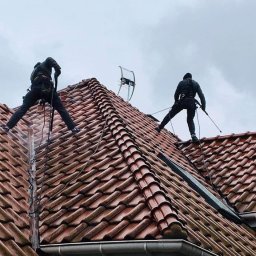 ALPINIX - Prace Wysokościowe Daniel Sadurski - Dwóch pracowników w uprzężach czyści dach z czerwonej dachówki w Radomiu. Widoczna antena i orynnowanie. Praca na wysokości, pochmurne niebo.