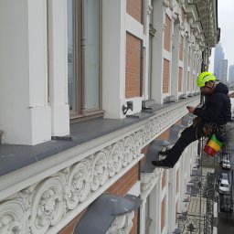 ALPINIX - Prace Wysokościowe Daniel Sadurski - Pracownik w kasku, na linach, kontroluje elewację budynku z dekoracyjnymi detalami. W tle widoczna panorama miasta z Pałacem Kultury.