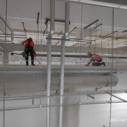 ALPINIX - Prace Wysokościowe Daniel Sadurski - Dwóch pracowników w uprzężach, odkurza i czyści konstrukcje stalowe pod sufitem w hali. Widoczne rury, kable i elementy mocujące. Praca na wysokości.