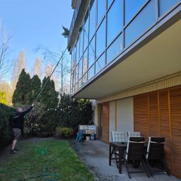 ALPINIX - Prace Wysokościowe Daniel Sadurski - Mężczyzna czyści szklaną fasadę budynku za pomocą długiej lancy z dyszą, stojąc na trawniku. Widoczne okna, zieleń i meble ogrodowe.