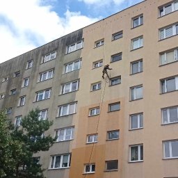 ALPINIX - Prace Wysokościowe Daniel Sadurski - Pracownik na linie czyści elewację bloku, widoczna różnica między umytą a brudną powierzchnią. Ujęcie z dołu, błękitne niebo w tle.