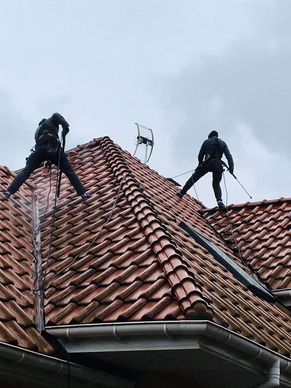 Dwóch pracowników w uprzężach czyści dach z czerwonej dachówki w Radomiu. Widoczna antena i orynnowanie. Praca na wysokości, pochmurne niebo.