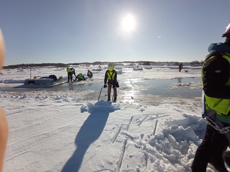 Grupa osób w odblaskowych ubraniach usuwa śnieg i lód z dachu pokrytego grubą warstwą śniegu w słoneczny dzień. Widoczne narzędzia i zabezpieczenia.