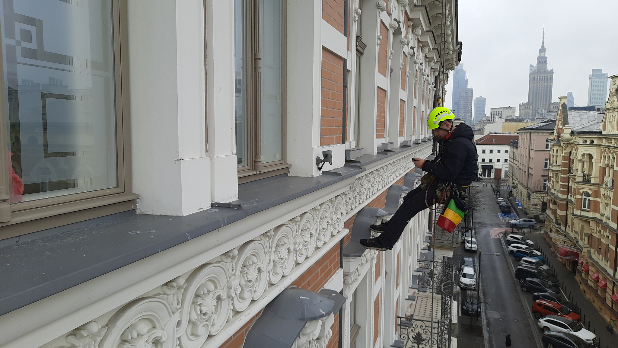 Pracownik w kasku, na linach, kontroluje elewację budynku z dekoracyjnymi detalami. W tle widoczna panorama miasta z Pałacem Kultury.