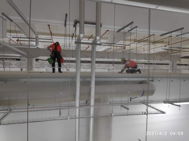 Dwóch pracowników w uprzężach, odkurza i czyści konstrukcje stalowe pod sufitem w hali. Widoczne rury, kable i elementy mocujące. Praca na wysokości.