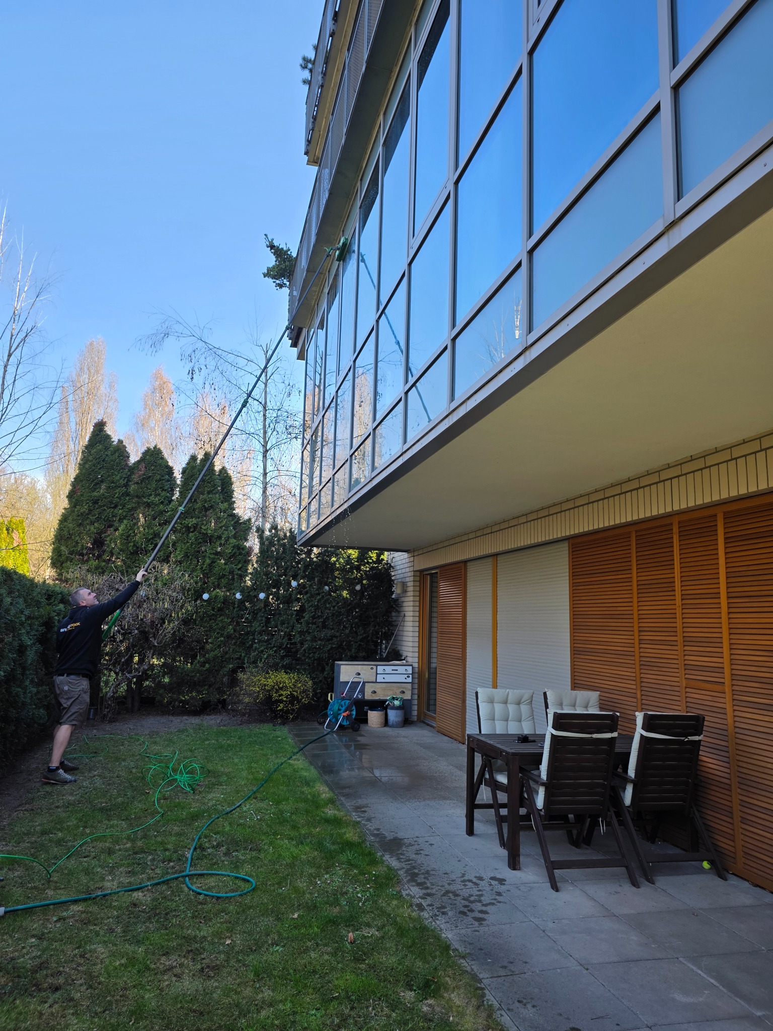 Mężczyzna czyści szklaną fasadę budynku za pomocą długiej lancy z dyszą, stojąc na trawniku. Widoczne okna, zieleń i meble ogrodowe.