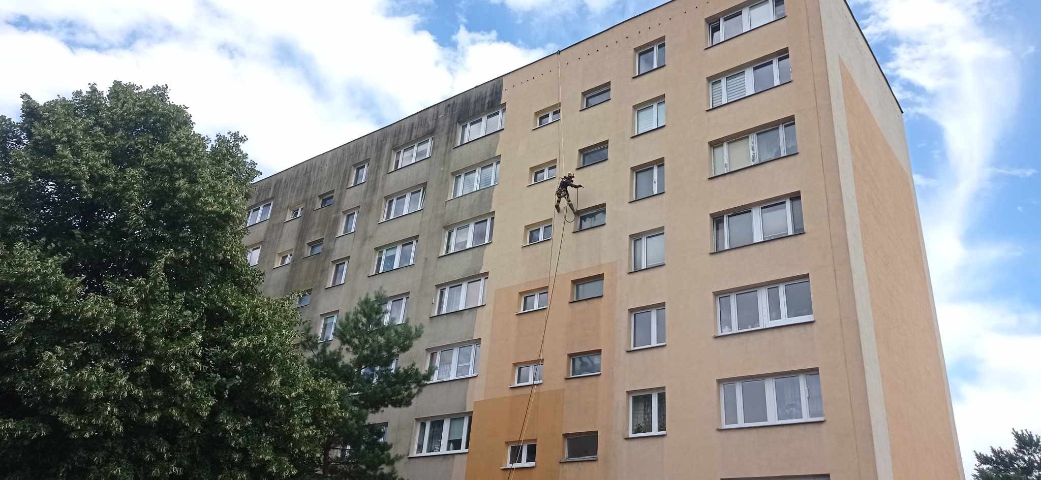 Pracownik na linie czyści elewację bloku, widoczna różnica między umytą a brudną powierzchnią. Ujęcie z dołu, błękitne niebo w tle.