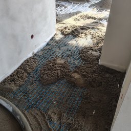 Gaw-Bud Posadzki maszynowe - Wnętrze w trakcie prac: na podłodze siatka zbrojeniowa, częściowo pokryta mokrą wylewką. W tle widoczne kopce materiału i rury ogrzewania podłogowego.