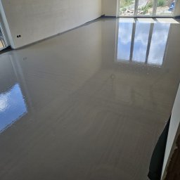 Gaw-Bud Posadzki maszynowe - Świeżo wylana, gładka posadzka w jasnym pomieszczeniu z dużymi oknami, odbijającymi niebo. Widoczne ślady po rozprowadzaniu masy, podkreślające ręczne wykonanie.
