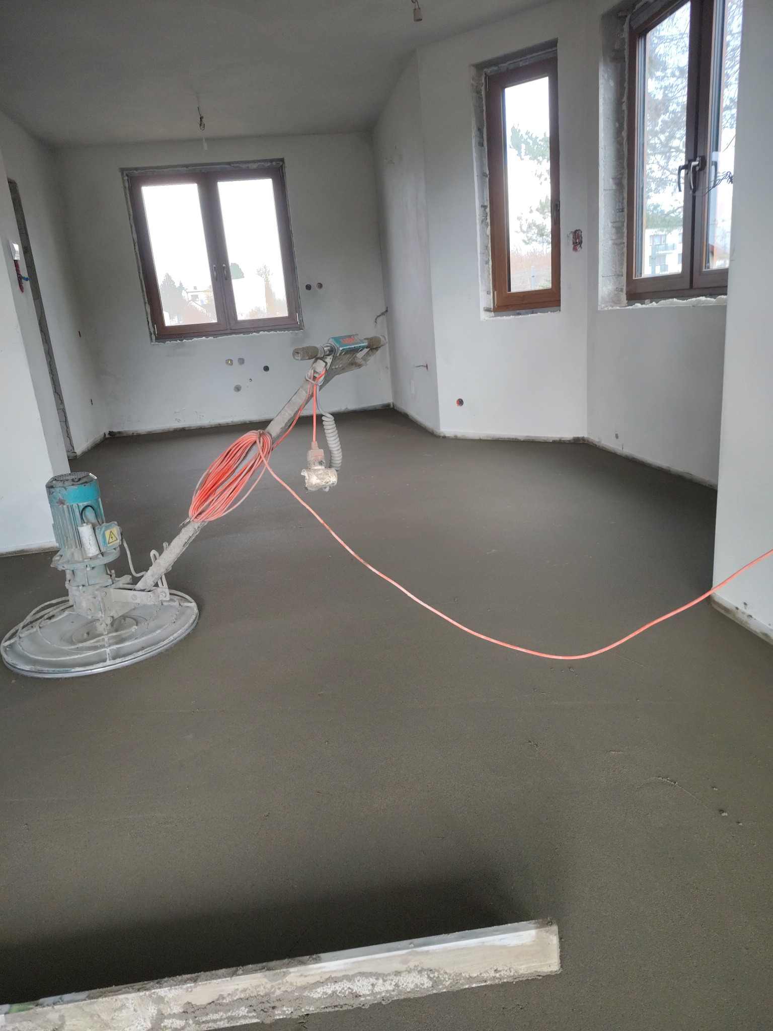 Wnętrze z maszyną do wygładzania wylewki, świeżo po wykonaniu prac. Widoczne okna i surowy stan ścian. Pomarańczowy kabel maszyny na szarej powierzchni.